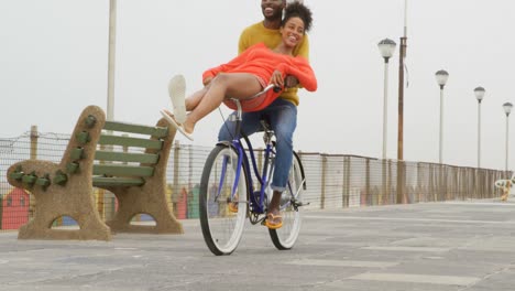 Front-view-of-young-black-couple-riding-bicycle-on-promenade-at-beach-on-a-sunny-day-4k