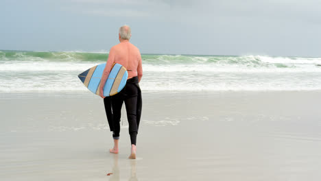 Rear-view-of-old-caucasian-man-walking-with-surfboard-at-beach-4k