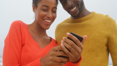 Front-view-of-young-black-couple-smiling-and-reviewing-photos-on-mobile-phone-at-beach-4k