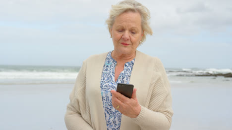 Vista-Frontal-De-Una-Anciana-Caucásica-Usando-Un-Teléfono-Móvil-En-La-Playa-4k