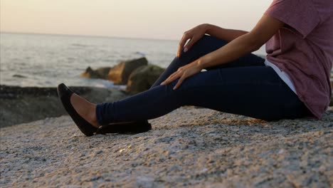 Thoughtful-mixed-race-woman-sitting-on-rock-at-beach-during-sunset-4k