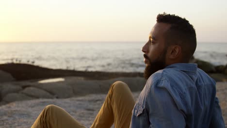 Thoughtful-mixed-race-man-sitting-on-rock-at-beach-during-sunset-4k