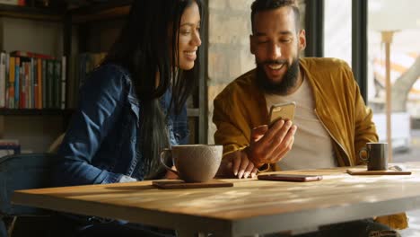 Front-view-of-happy-young-mixed-race-couple-discussing-over-mobile-phone-at-table-in-cafe-4k