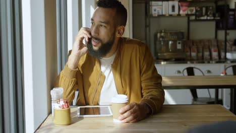 Front-view-of-happy-young-mixed-race-man-talking-on-mobile-phone-in-cafeteria-4k