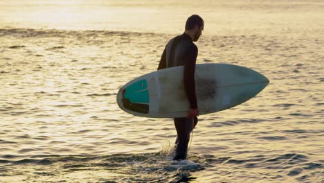 Side-view-of-mid-adult-caucasian-male-surfer-with-surfboard-walking-in-sea-during-sunset-4k