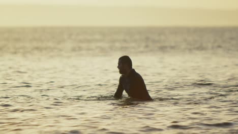 Side-view-of-mid-adult-caucasian-male-surfer-sitting-over-surfboard-in-sea-during-sunset-4k