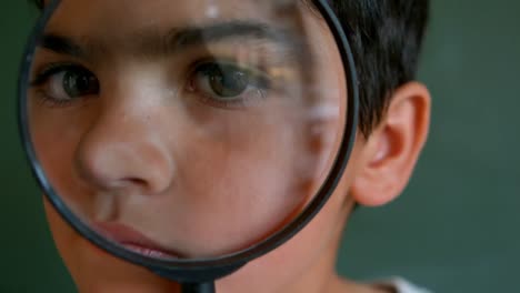 Close-up-of-Caucasian-schoolboy-looking-through-magnifying-glass-against-green-board-in-classroom-4k