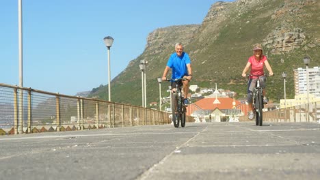 Front-view-of-active-senior-Caucasian-couple-riding-bicycle-on-a-promenade-at-the-beach-4k-