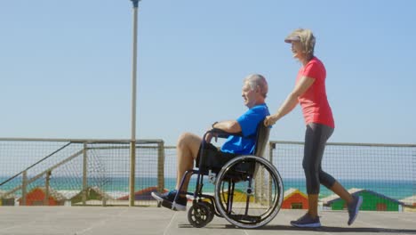 Side-view-of-active-senior-Caucasian-woman-pushing-senior-man-on-wheelchair-at-beach-4k