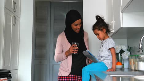 Young-mother-with-a-hijab-drinking-a-glass-of-water-near-her-daughter-in-the-kitchen-4k