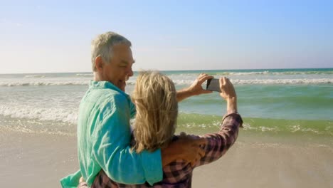 Rückansicht-Eines-Aktiven-älteren-Kaukasischen-Paares,-Das-Am-Strand-Ein-Selfie-Mit-Dem-Mobiltelefon-Macht,-4k