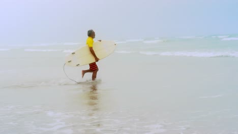 Vista-Lateral-De-Un-Surfista-Afroamericano-Senior-Activo-Corriendo-Hacia-El-Mar-En-La-Playa-4k