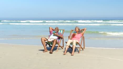 Front-view-of-active-senior-African-American-couple-having-drinks-on-deckchair-at-beach-4k