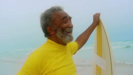 Vista-Frontal-De-Un-Feliz-Hombre-Afroamericano-Senior-Activo-Con-Tabla-De-Surf-De-Pie-En-La-Playa-4k
