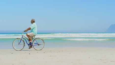 Side-view-of-active-senior-African-American-man-riding-bicycle-on-beach-in-the-sunshine-4k