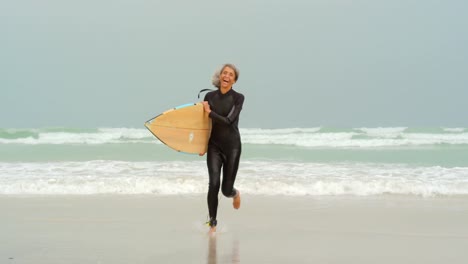 Front-view-of-active-senior-African-American-female-surfer-with-surfboard-running-on-the-beach-4k