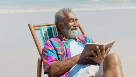 Vorderansicht-Eines-Aktiven-älteren-Afroamerikanischen-Mannes,-Der-Auf-Einem-Liegestuhl-Am-Strand-Ein-Buch-Liest,-4k