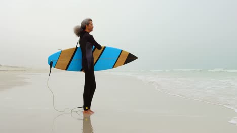 Side-view-of-active-senior-African-American-female-surfer-with-surfboard-standing-on-the-beach-4k