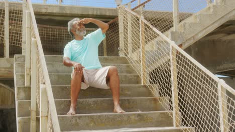 Front-view-of-active-senior-African-American-man-with-shielding-eye-sitting-on-stair-at-promenade-4k