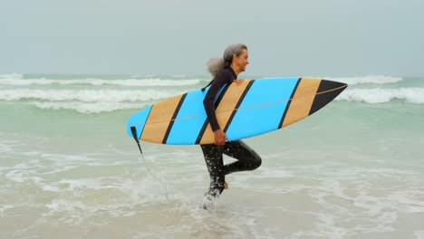 Side-view-of-active-senior-African-American-female-surfer-running-towards-sea-on-the-beach-4k