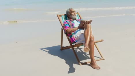 Vorderansicht-Eines-Aktiven-älteren-Afroamerikanischen-Mannes,-Der-Sich-Auf-Einem-Liegestuhl-Mit-Einem-Buch-Am-Strand-Entspannt,-4k
