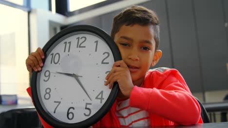 Front-view-of-Asian-schoolboy-holding-wall-clock-at-desk-in-classroom-at-school-4k