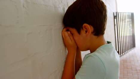 Side-view-of-Caucasian-schoolboy-covering-his-face-with-hands-while-leaning-on-wall-in-corridor-4k