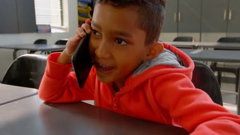 Front-view-of-Asian-schoolboy-talking-on-mobile-phone-at-desk-in-classroom-at-school-4k