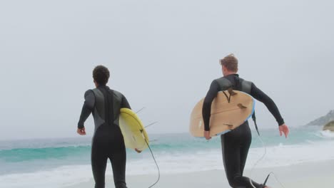 Rückansicht-Von-Zwei-Männlichen-Surfern,-Die-Zusammen-Mit-Einem-Surfbrett-Am-Strand-Laufen,-4k