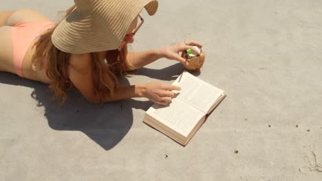Vista-De-ángulo-Alto-De-Una-Mujer-Caucásica-Con-Sombrero-Leyendo-Un-Libro-En-La-Playa-4k