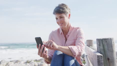 Caucasian-woman-enjoying-free-time-by-sea-on-sunny-day-using-smartphone