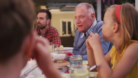 Senior-man-taking-to-family-members-while-having-dinner-outdoors