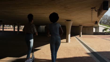 Two-mixed-race-women-riding-electric-scooter-under-bridge