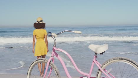 Afroamerikanische-Frau,-Die-Mit-Einem-Fahrrad-Im-Vordergrund-Am-Meer-Spaziert