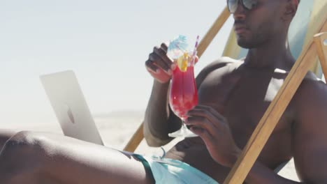 African-american-man-using-his-laptop-at-beach-and-drinking-juice-