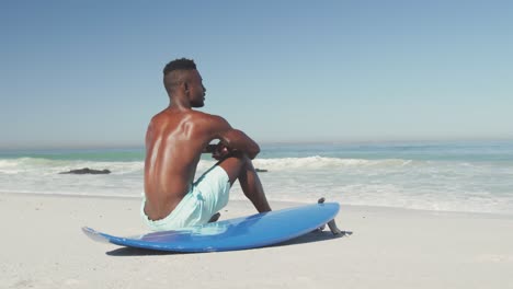 African-American-man-sitting-at-beach-with-surfboard