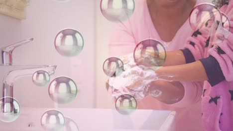 Animation-of-bubbles-floating-up-over-a-girl-and-her-grandmother-washing-their-hands-together-