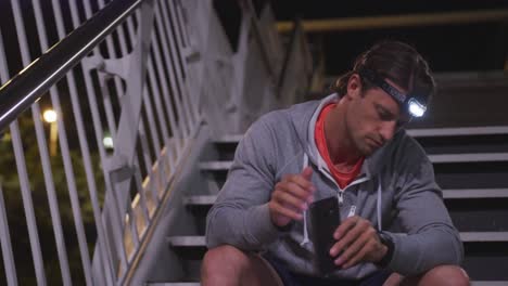 Caucasian-male-wearing-a-headlamp,-checking-his-smartwatch,-using-his-phone-on-stairs-in-the-evening