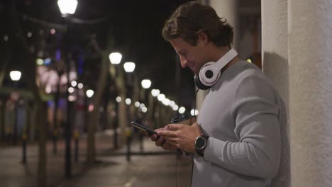 Caucasian-male-wearing-headphones,-drinking,-using-his-phone-in-the-evening