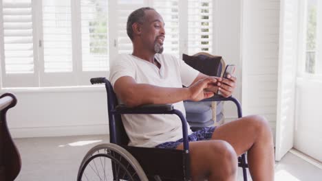 Älterer-Afroamerikanischer-Mann-Im-Rollstuhl,-Der-Sein-Telefon-Benutzt-Und-Zu-Hause-Lächelt