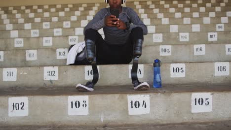 Behinderter-Mischlingsmann-Mit-Beinprothesen,-Der-Auf-Einem-Stadion-Sitzt-Und-Ein-Smartphone-Benutzt