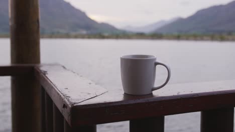 A-view-of-a-lake-and-mountains-during-sunset-on-a-cloudy-day-seen-from-log-cabin-window-with-a-mug-i