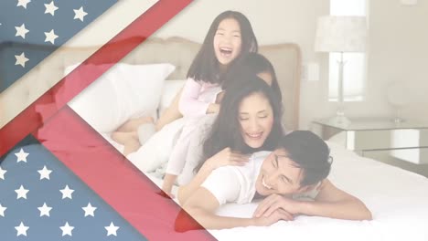 Animation-of-U.S.-flag-over-Asian-family-playing-on-bed