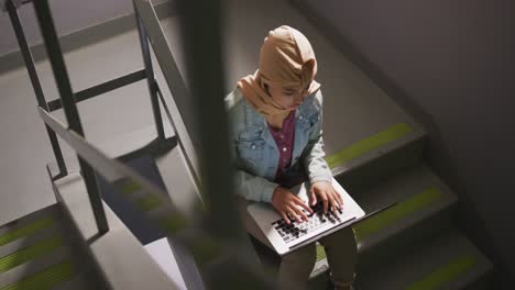 Asian-female-student-wearing-a-beige-hijab-sitting-on-stairs-and-using-a-laptop