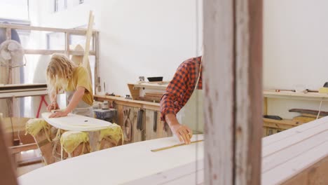 Two-Caucasian-male-surfboard-makers-working-in-their-studio-and-making-a-wooden-surfboard-together