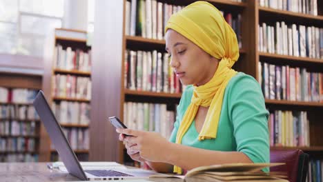 Eine-Asiatische-Studentin,-Die-Einen-Gelben-Hijab-Trägt,-Sitzt-An-Einem-Schreibtisch-Und-Benutzt-Einen-Laptop-Und-Ein-Smartphone