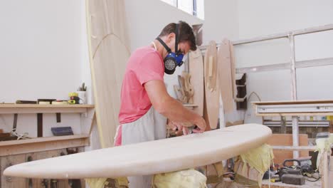 Caucasian-male-surfboard-maker-wearing-a-face-mask-and-preparing-to-polishing-a-surfboard