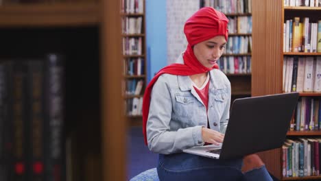 Una-Estudiante-Asiática-Con-Un-Hijab-Rojo-Estudiando-En-Una-Biblioteca-Y-Usando-Una-Computadora-Portátil