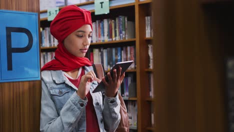 An-Asian-female-student-wearing-a-red-hijab-studying-in-a-library-and-using-tablet