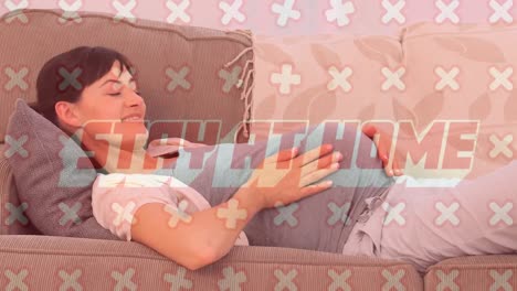 Animation-of-text-Stay-At-Home-with-crosses-moving-over-pregnant-Caucasian-woman-lying-on-a-couch-an